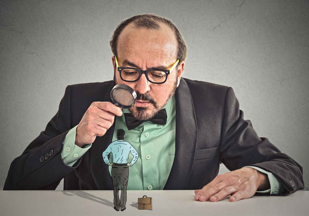 Curious corporate businessman skeptically meeting looking at small employee standing on table through magnifying glass isolated office grey wall background. Human face expression, attitude, perception.jpeg